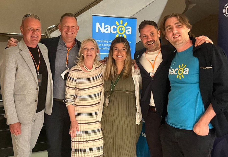Six people standing together in front of a Nacoa banner at an event, two men wearing lanyards.